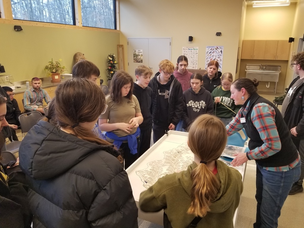 Group of students gathered around a table with a large map, listening to an instructor explaining details in a classroom setting.