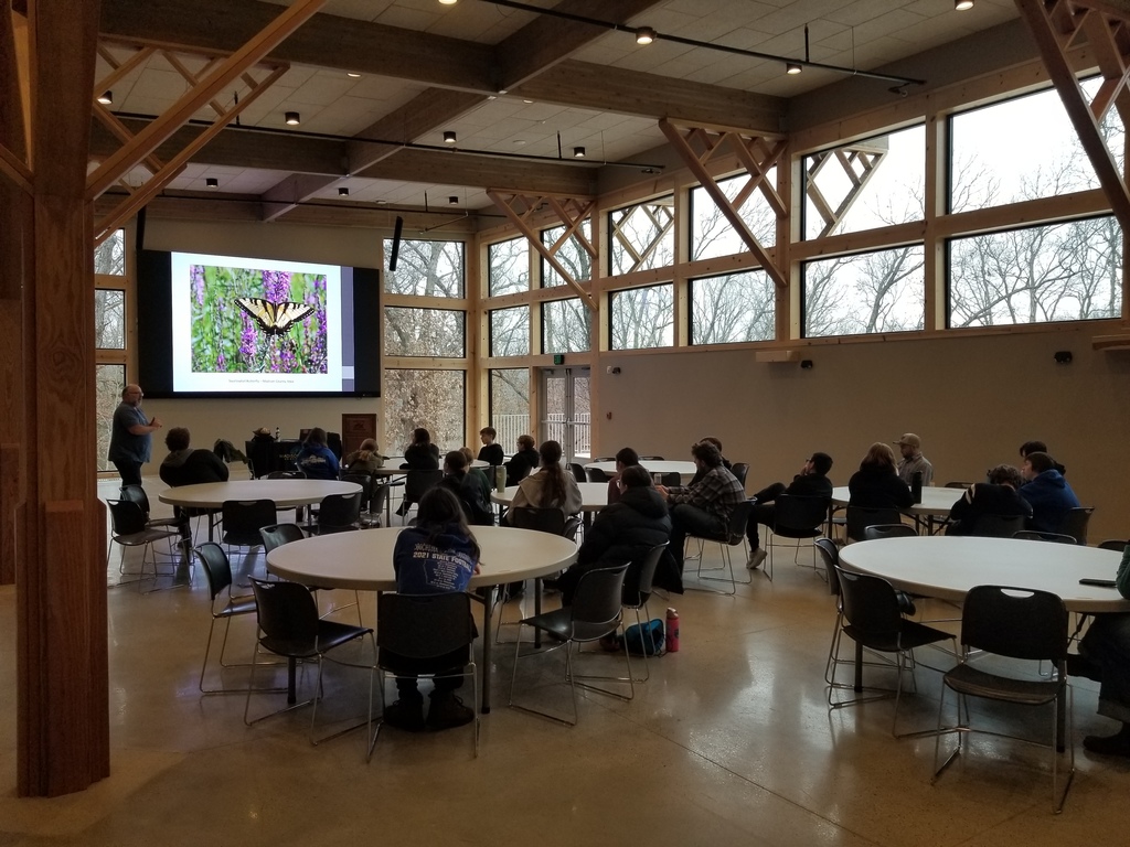 A group of people seated at round tables in a spacious room watching a presentation on a large screen showing a butterfly image.