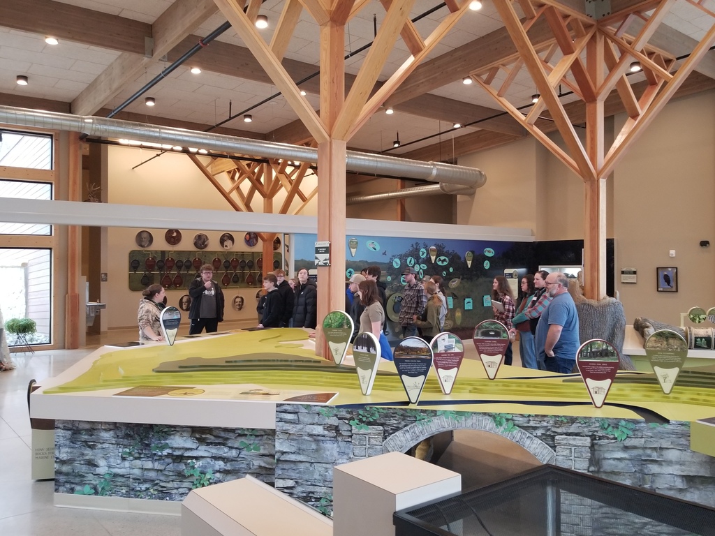 Group of people gathered around a large interactive exhibit table inside a spacious, modern building with wooden beams and high ceilings.