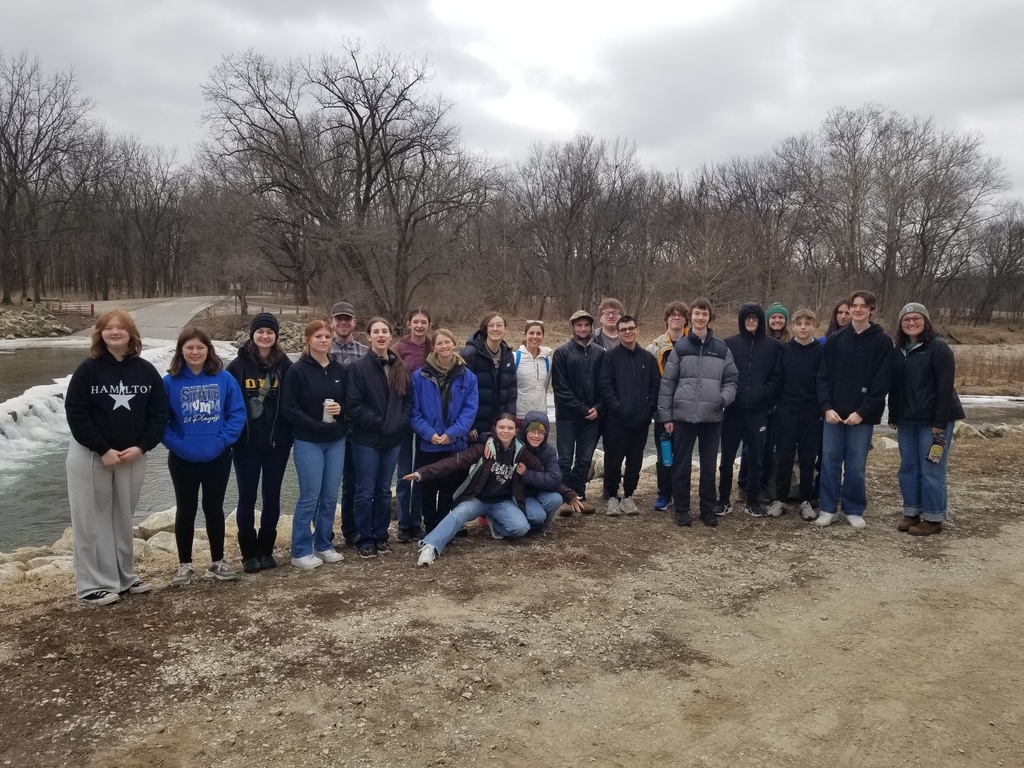 Group of young adults and a few older individuals posing outdoors on a cloudy day near a river and leafless trees.