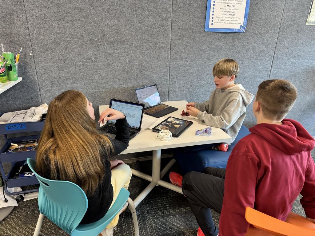 Three students collaborate at a white table with laptops and tablets in a classroom setting.