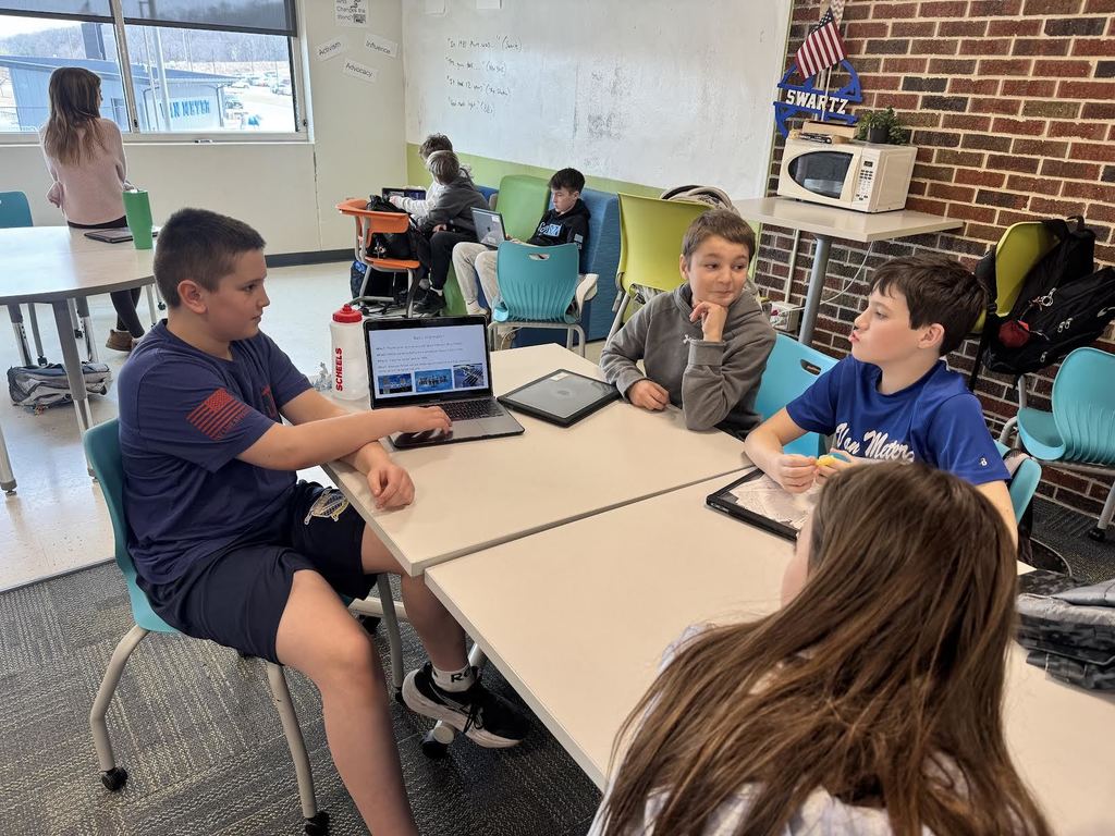 A group of students sitting around tables in a classroom, engaged with laptops and tablets during a collaborative activity.
