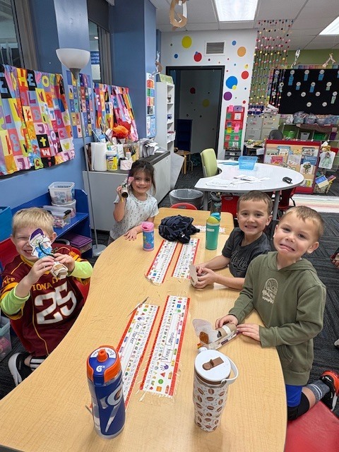 Four children sitting around a classroom table, drinking from water bottles and eating snacks during a break.