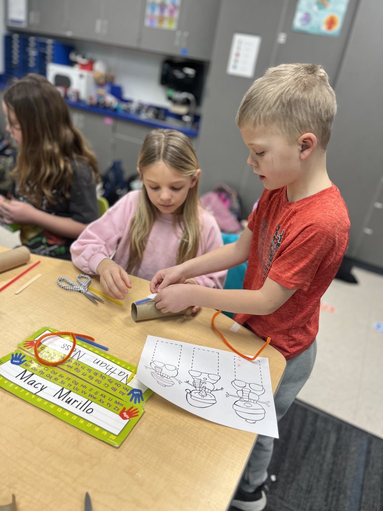 Two children working together on a craft project using a cardboard tube and pipe cleaners at a classroom table.