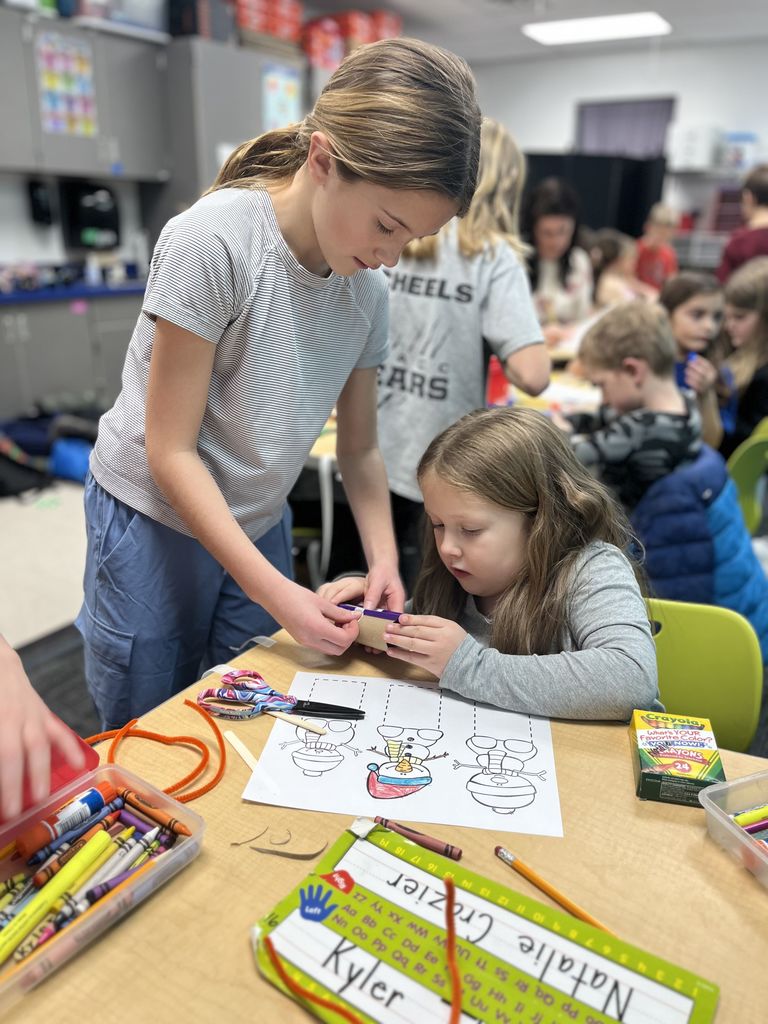 Two children collaborate on a colorful drawing activity at a classroom table filled with crayons and art supplies.