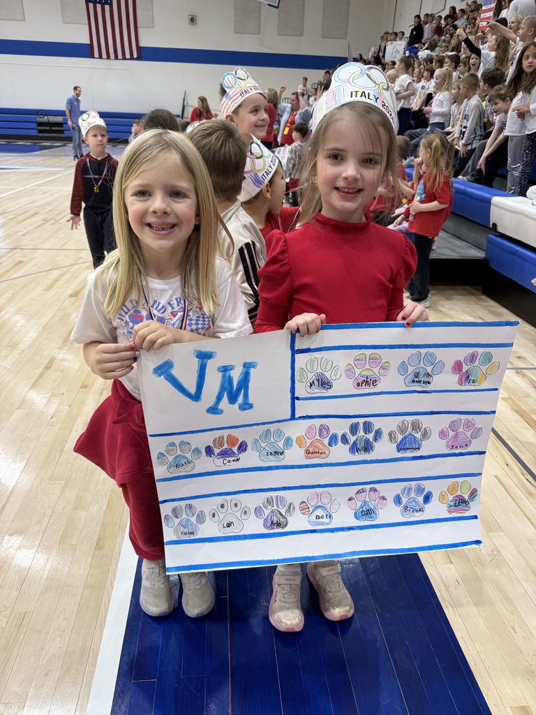 Two children in a gym holding a colorful poster with paw prints and the letters 'VM' during a school event.