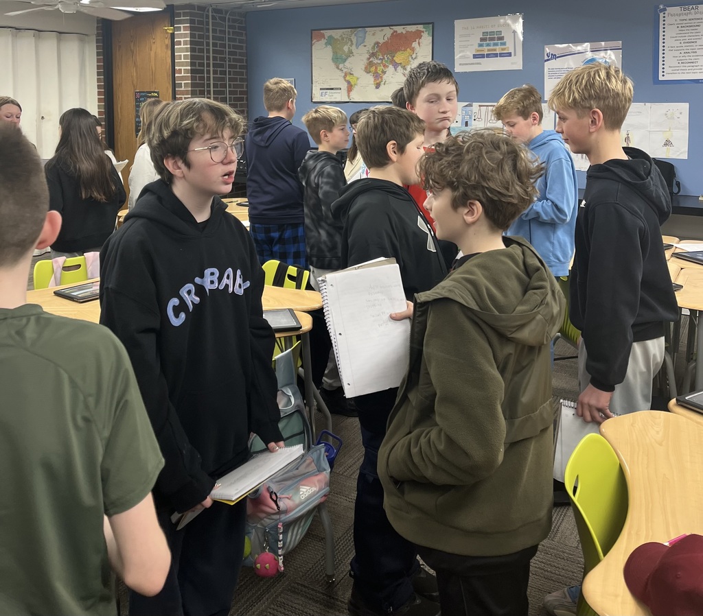 Students stand in small groups, talking and holding notebooks in a classroom with desks, a world map, and posters on blue walls.