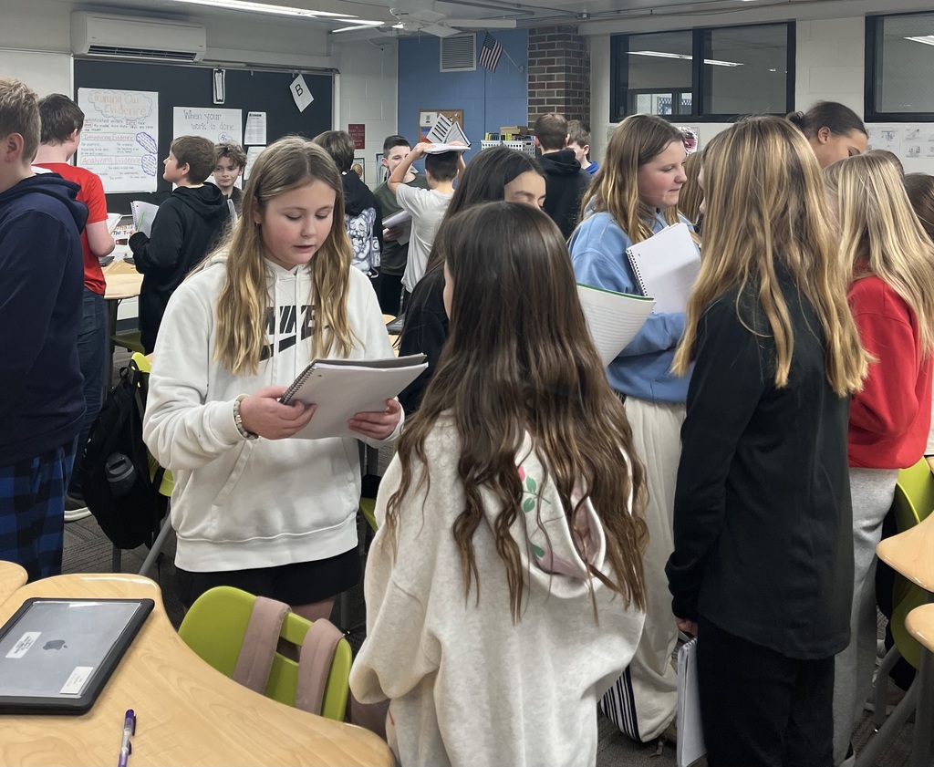 Students stand in small groups, reading from notebooks and discussing in a busy classroom with posters and a whiteboard in the background.
