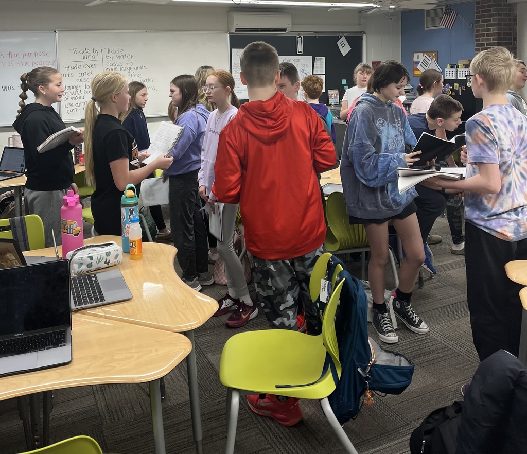 Students stand in small groups, reading from notebooks and discussing in a classroom with desks, laptops, and a whiteboard behind them.