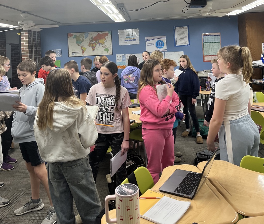Students stand in small groups around desks, discussing and holding notebooks in a busy classroom with maps and posters on blue walls.