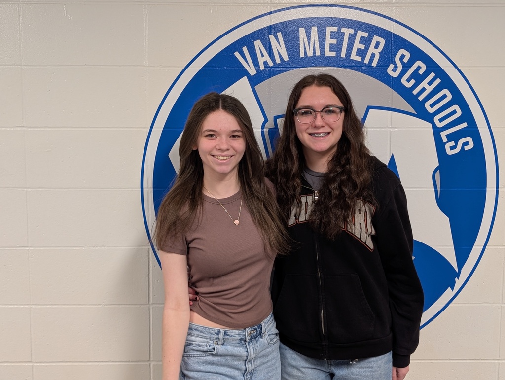 Two teenage girls standing in front of a Van Meter Schools logo on a beige wall, smiling and posing together.