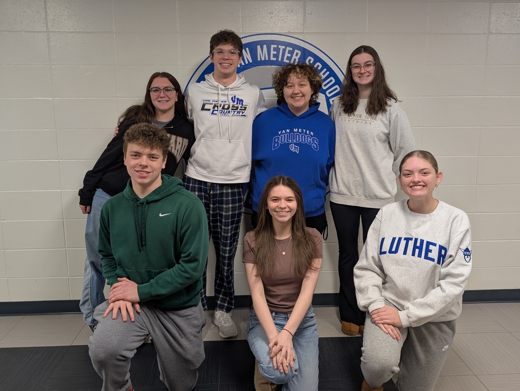 Seven students posing in front of a school emblem on a beige wall inside a hallway.