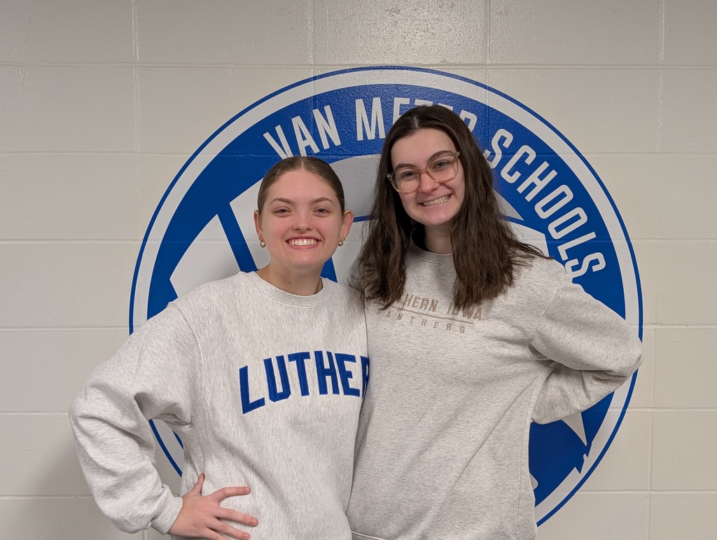 Two young women stand side by side in front of a Van Meter Schools logo on a white wall, both wearing light gray sweatshirts with school names.