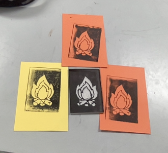 Four handmade prints of a stylized campfire design on colored paper, arranged on a gray surface.