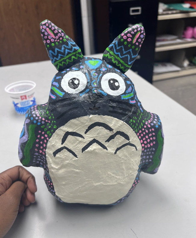Hand holding a colorful, hand-painted Totoro figurine with intricate patterns in blue, green, pink, and purple on a white table.