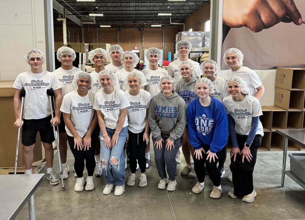 Group of young people wearing matching white shirts posing inside a warehouse with shelves and boxes in the background.