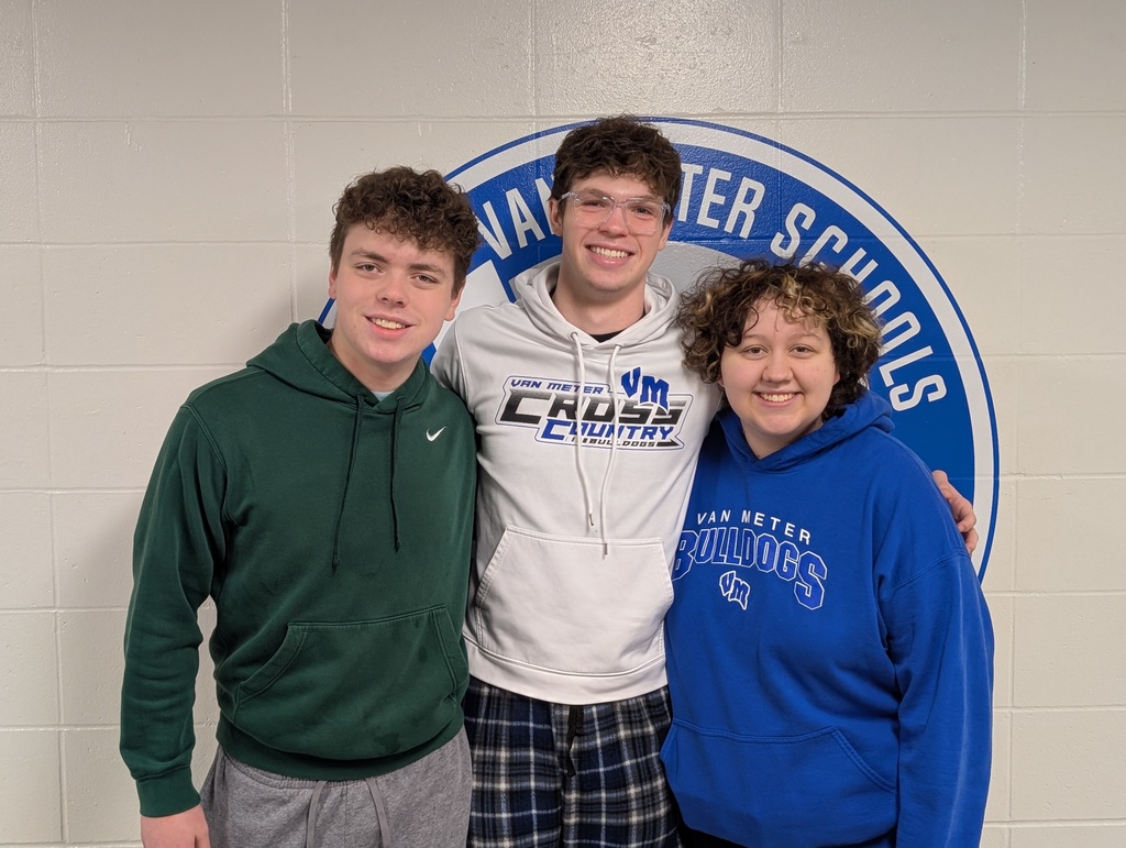 Three teenage students standing together in front of a Van Meter Schools emblem on a beige wall, wearing casual hoodies and smiling.