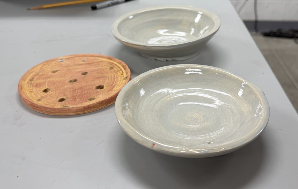 Two handmade ceramic bowls with a glossy light gray glaze sit on a table next to a round wooden disc with holes.