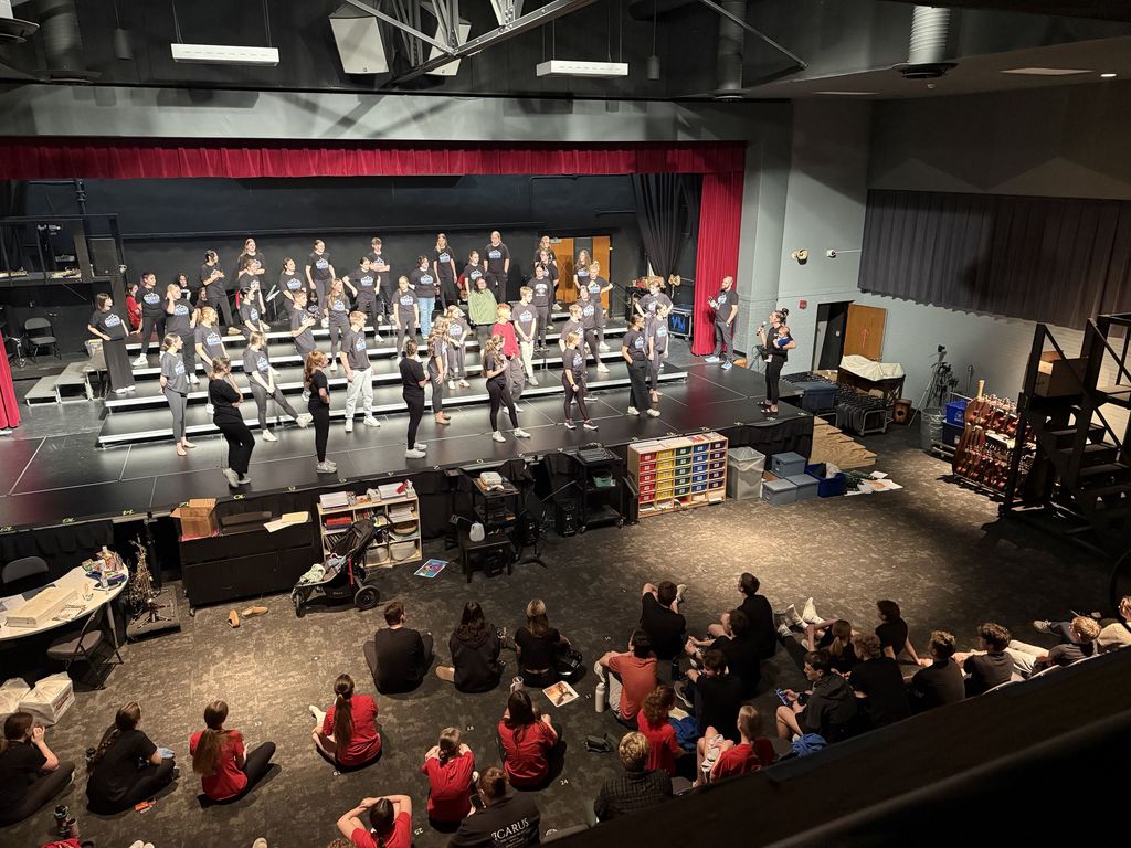 A large group of performers rehearsing on a theater stage with an audience seated on the floor watching attentively.