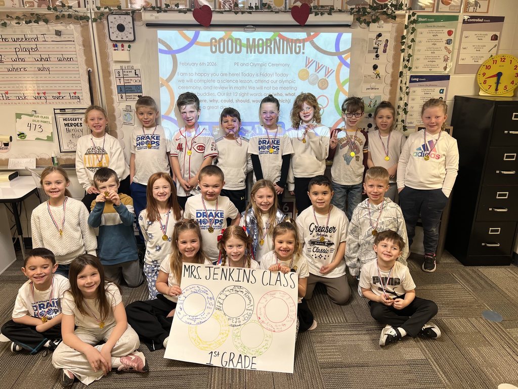 Group of first-grade students and their teacher posing in a classroom holding a sign that reads 'Mrs. Jenkins Class 1st Grade'.