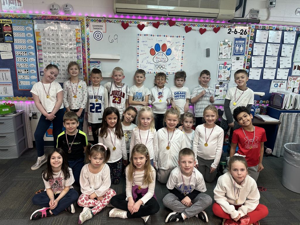 Group of young students posing in a colorful classroom with educational posters and a whiteboard behind them.