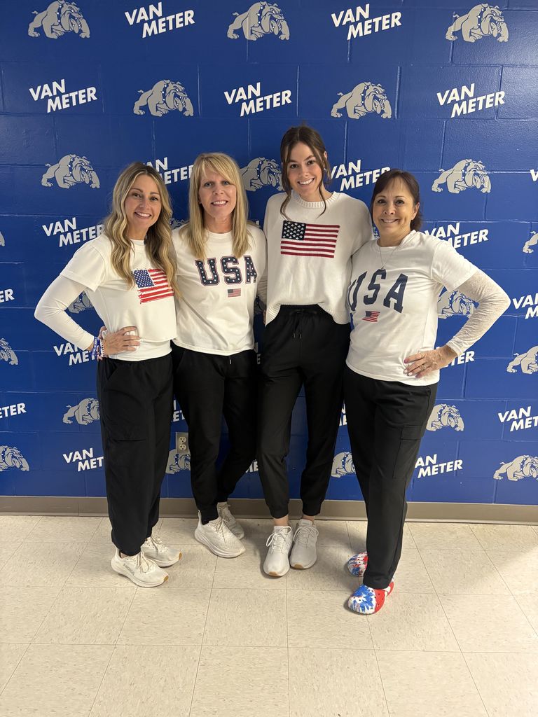Four women standing in front of a blue Van Meter wall, wearing white USA-themed shirts and black pants, smiling together.