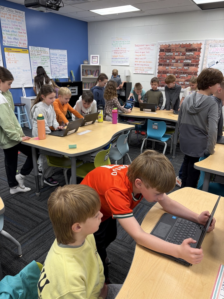 Elementary students collaborating on laptops in a colorful classroom with educational posters on the walls.
