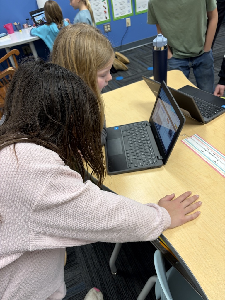 Two students closely collaborating on a laptop at a classroom table with other children and educational posters in the background.