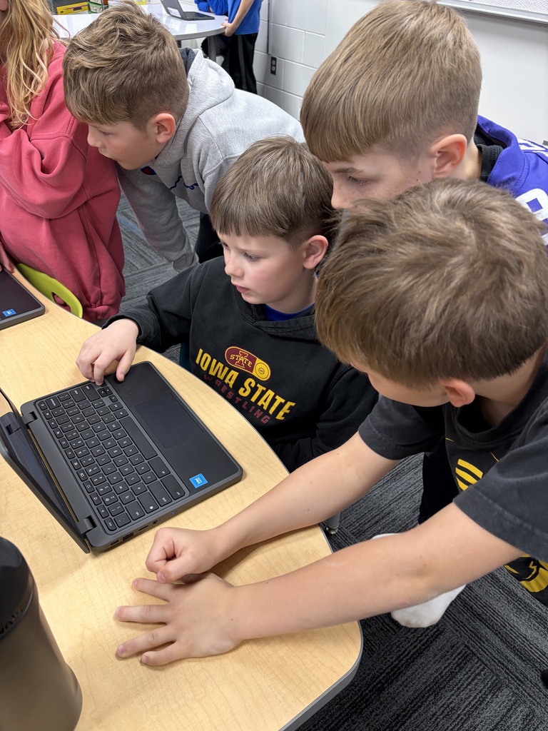 Four young boys closely gathered around a laptop on a classroom desk, focused on the screen during a group activity.