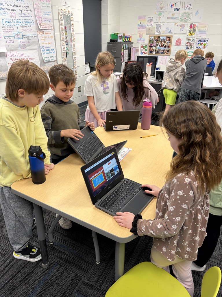 Elementary students collaborating around a table using laptops in a colorful classroom setting.