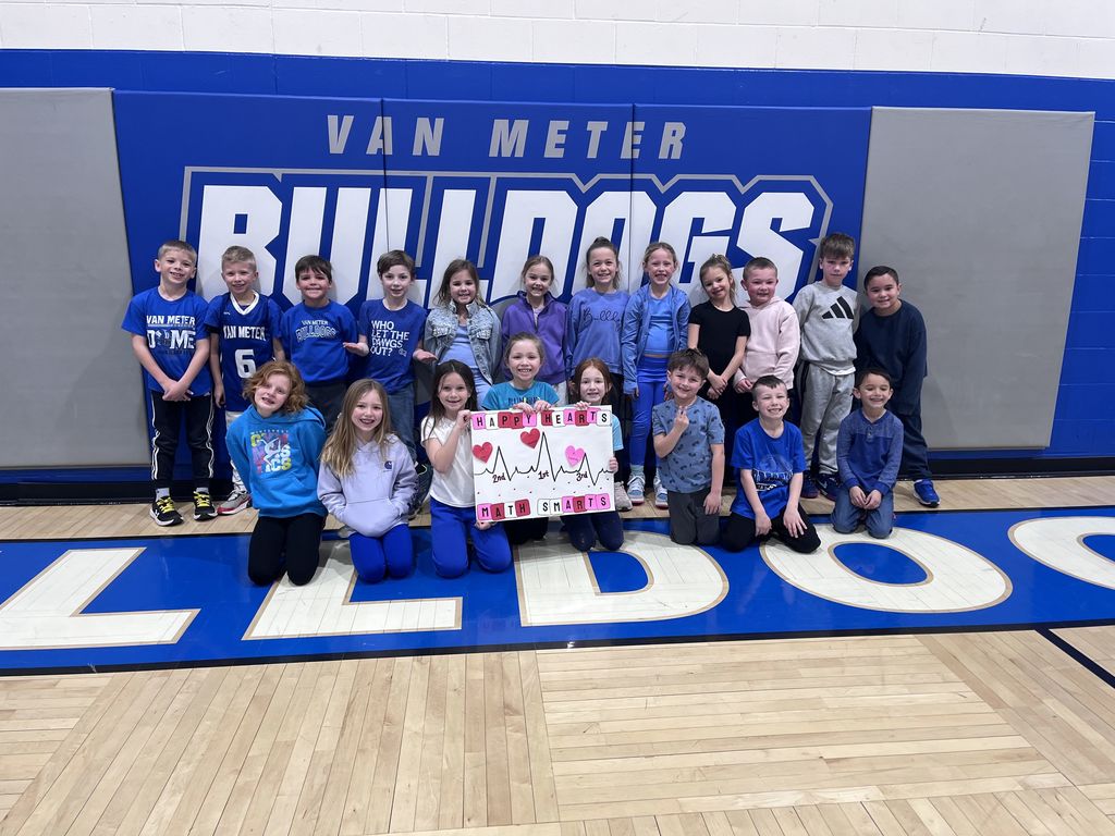 Group of elementary school children posing in front of a Van Meter Bulldogs gym wall holding a colorful handmade poster.