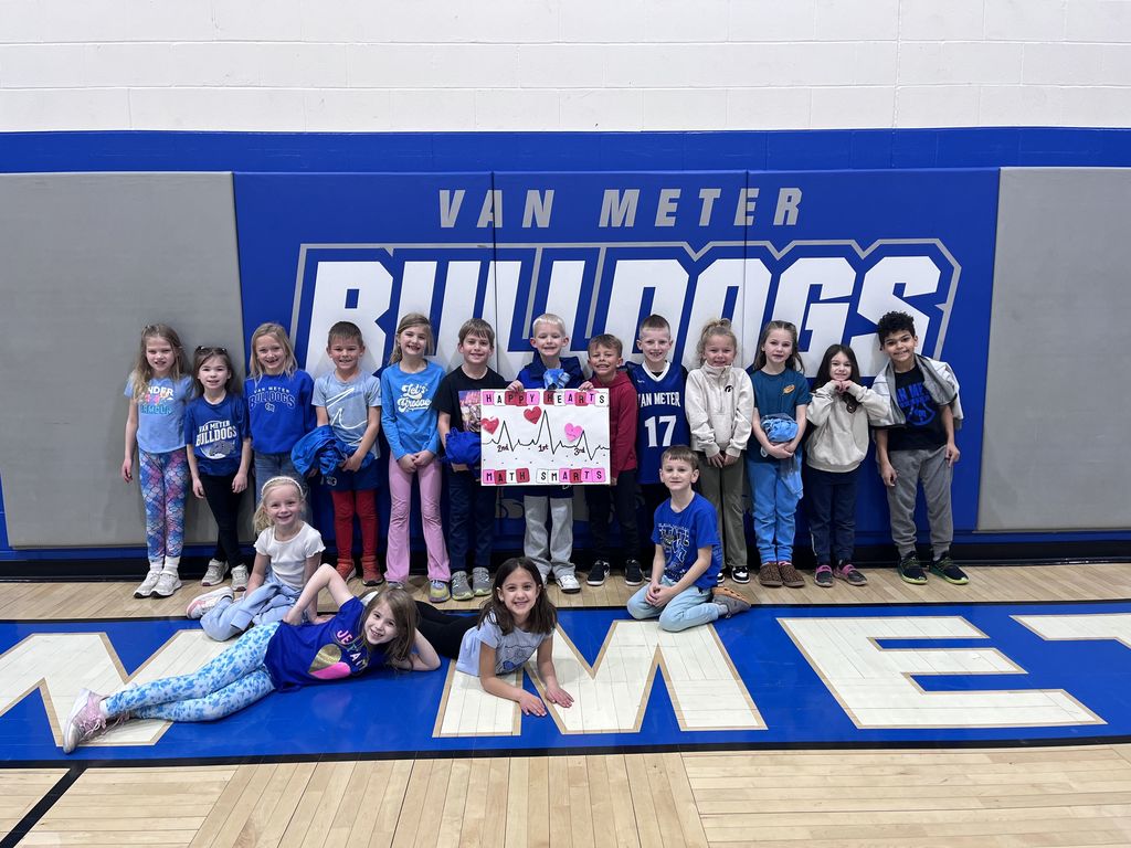 Group of young children posing in front of a Van Meter Bulldogs gym wall holding a colorful poster with heart drawings.