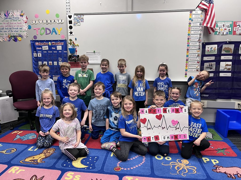 Group of young students in a classroom sitting and standing on a colorful alphabet rug, holding a poster that reads 'Happy Hearts Math Smarts' with heart and heartbeat illustrations.