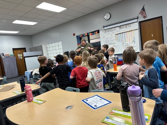 Teacher stands at front of classroom leading activity as young students gather around desks and watch attentively.