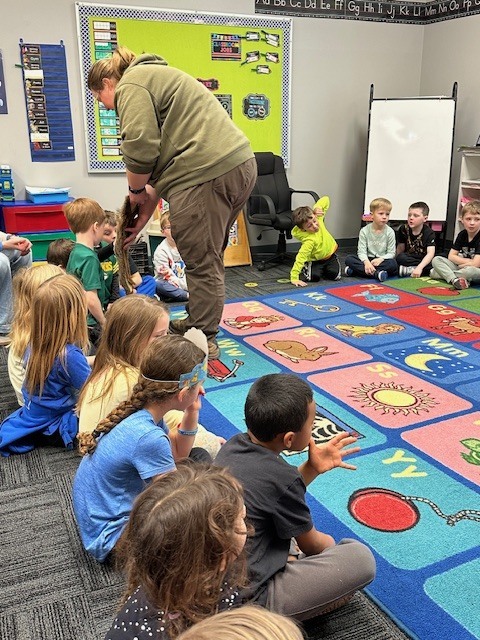 Teacher engaging a group of young children seated on a colorful alphabet and weather-themed carpet in a classroom setting.