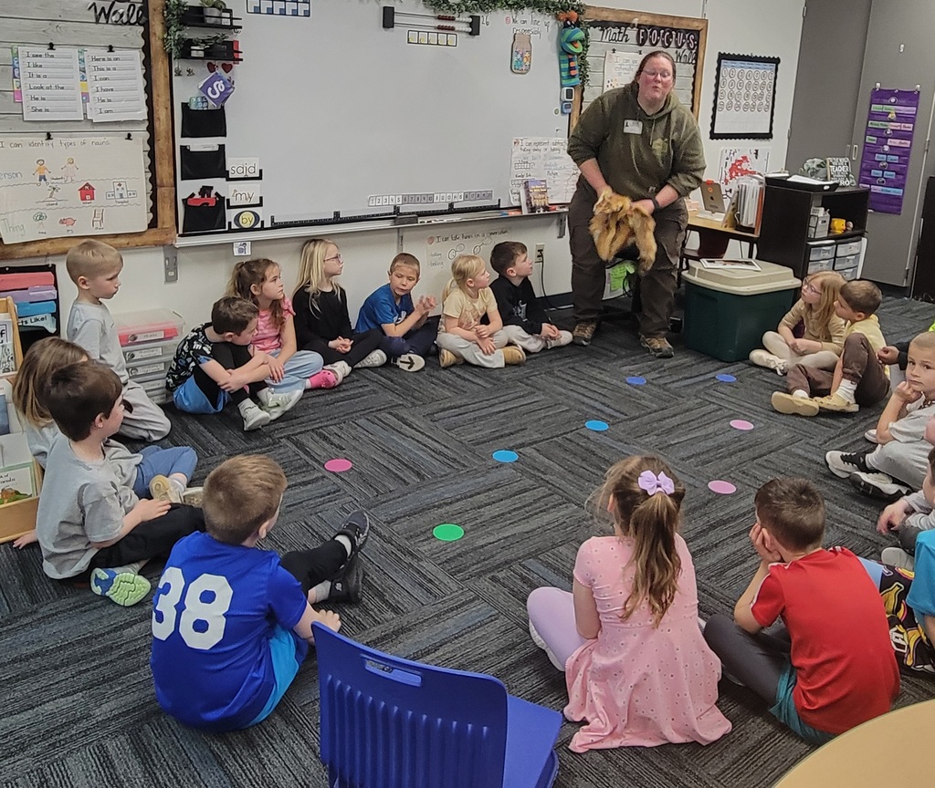 A group of young children sitting in a classroom circle attentively watching a man holding a small dog during a lesson.