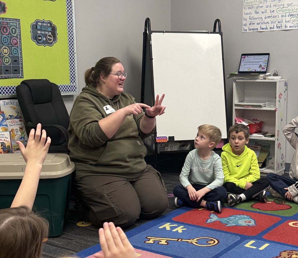 Teacher kneels on classroom rug, counting on fingers as young children sit and raise hands during lesson.