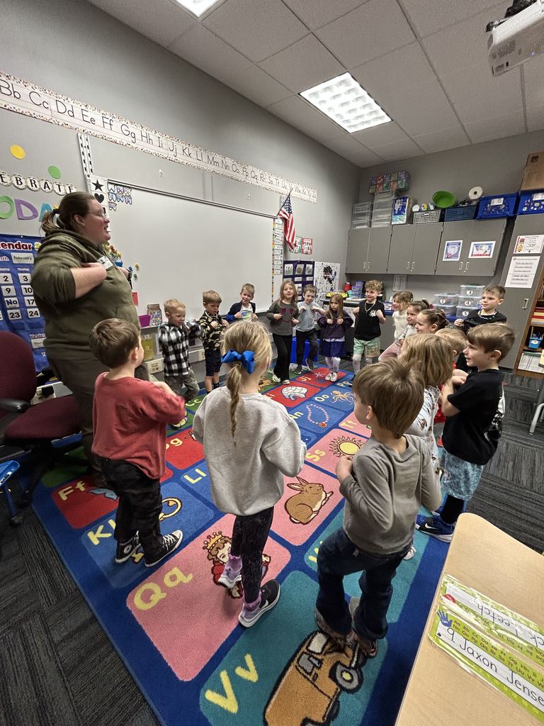Young children standing in a circle on a colorful alphabet rug during a classroom activity led by a teacher.
