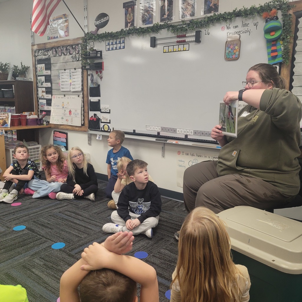 Teacher reading a book aloud to young students seated on a classroom carpet with colorful dots.