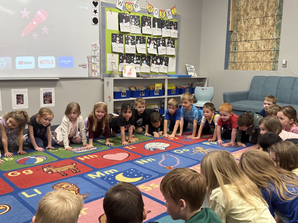 Children kneel in a circle on colorful alphabet rug, leaning in for group activity in classroom.