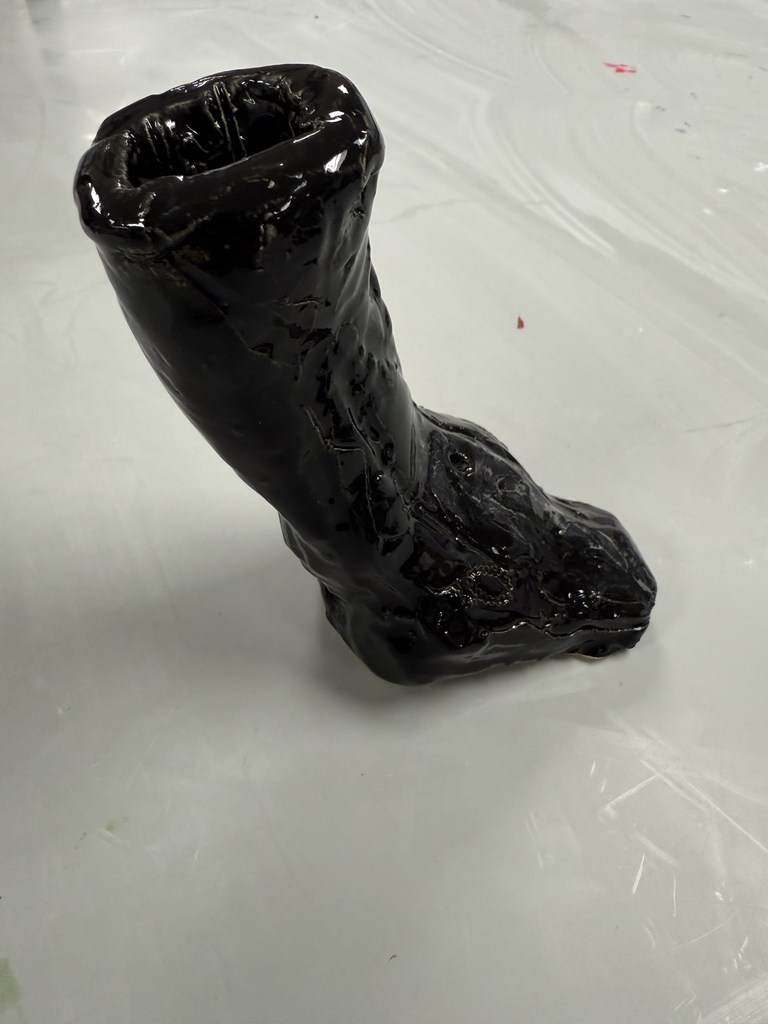 Glossy black ceramic sculpture shaped like a boot standing upright on a white surface.