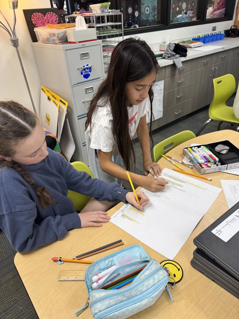 Two girls collaborate on a drawing project at a classroom table filled with pencils, markers, and paper.
