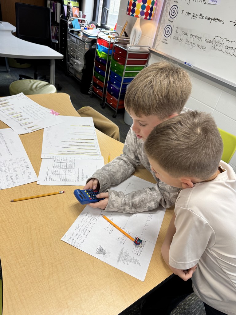 Two young boys collaborate on math homework using a calculator at a classroom desk filled with papers and pencils.