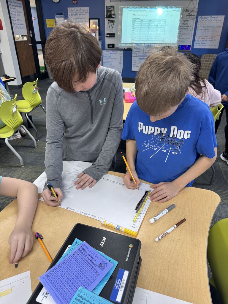 Two boys collaborate on a worksheet at a classroom desk filled with school supplies and a laptop.