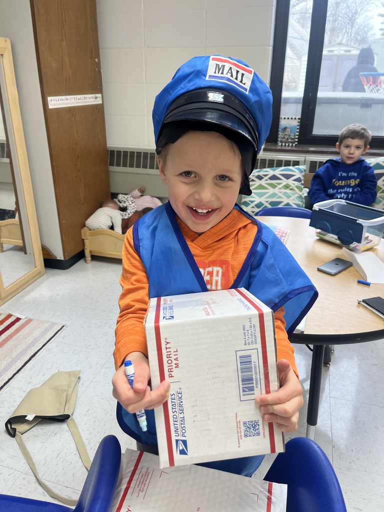 Child dressed as a mail carrier holding a USPS Priority Mail box in a classroom setting.