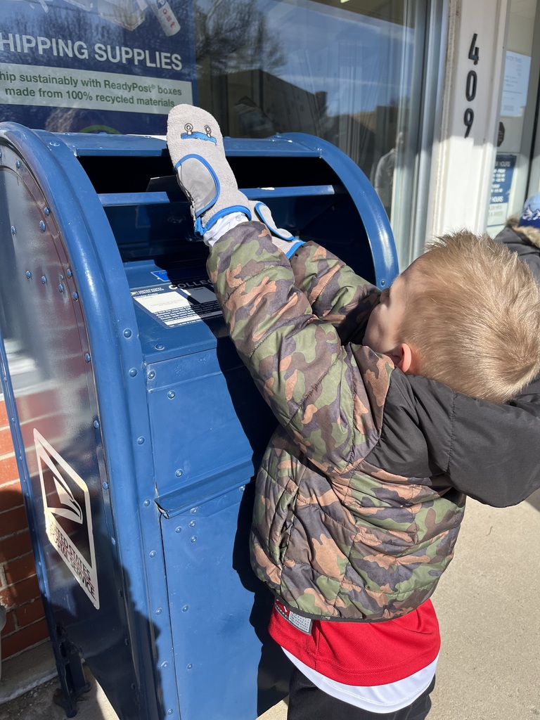 Child in winter jacket and patterned hat reaches up to mail a letter in a blue USPS mailbox while an adult steadies it.