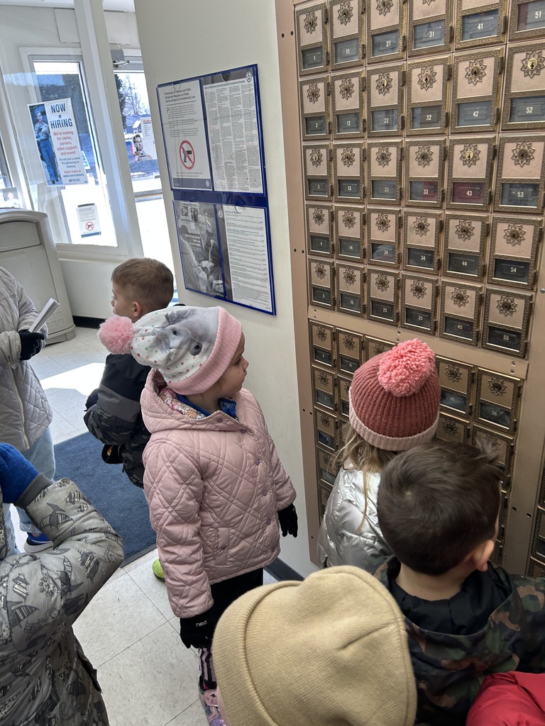 Children wearing winter coats and hats gather around a wall of vintage postal mailboxes inside a post office lobby.