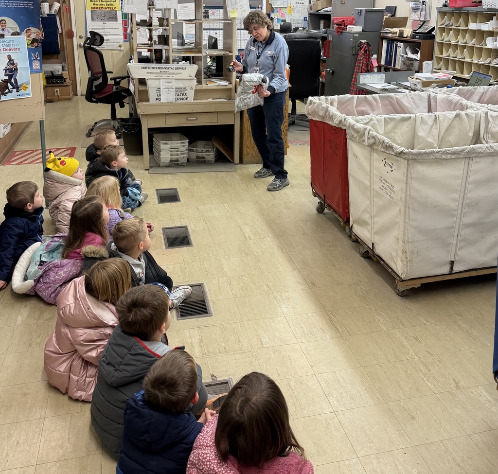 Postal worker shows a package scanner to a group of young children seated on the floor during a behind-the-scenes post office tour.