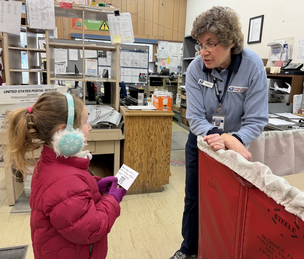Postal worker leans on a mail bin while speaking with a young girl in a red coat and earmuffs holding a letter inside a busy post office.