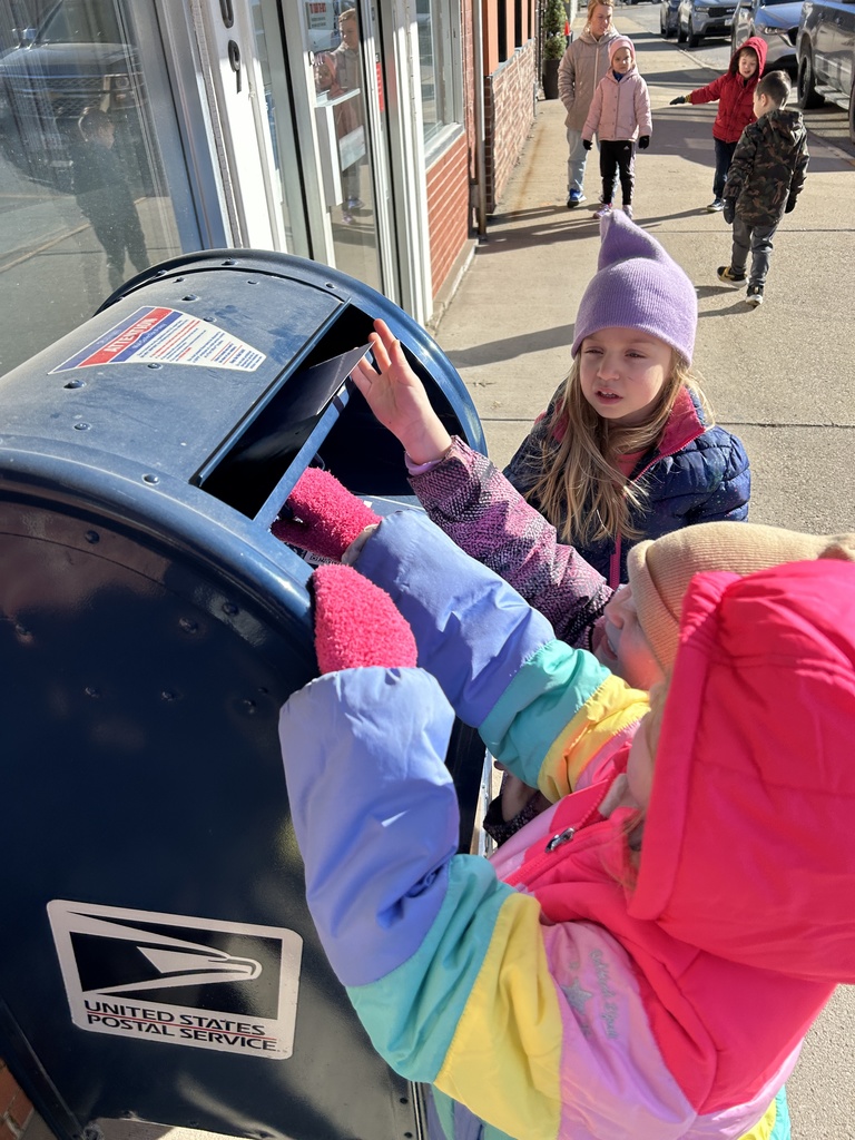 Two children in colorful winter jackets mailing a letter at a blue USPS mailbox on a sunny sidewalk.